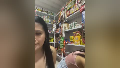 Video of a Latina woman with long black hair and a medium complexion, wearing a sleeveless top, in a cluttered supermarket aisle filled with assorted packaged goods.