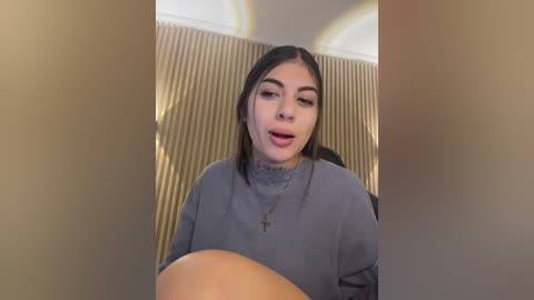 A video of a young woman with long dark hair and a gray sweater, making a surprised face, standing in front of a beige, vertically-striped wall.