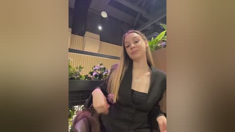 Video of a young woman with long, straight brown hair, wearing a black blazer and black top, standing in a modern indoor setting with wooden panels and green plants.