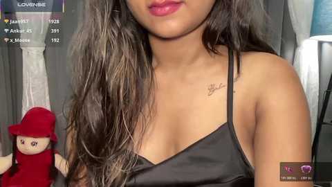 Media: A video of a young woman with medium brown skin and long, wavy dark hair, wearing a black satin camisole. She has a small tattoo on her collarbone. The background includes a red dress and a white curtain.
