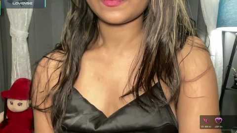 A close-up video of a young woman with long, dark hair wearing a black satin dress, partially visible, in a dimly lit room with a red cushion and white curtains in the background.