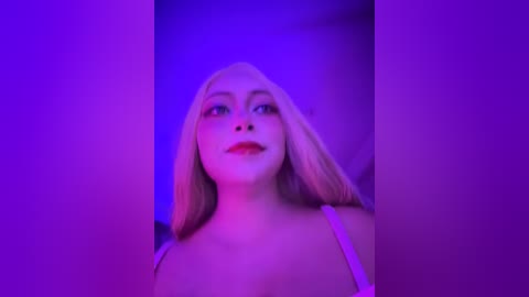 A video of a blonde woman with fair skin and red lipstick, wearing a white spaghetti strap top, against a purple background.