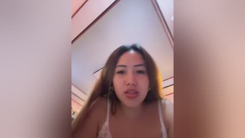 A blurry video of an Asian woman with long brown hair, wearing a white lace bra, standing in a dimly lit room with a triangular ceiling.