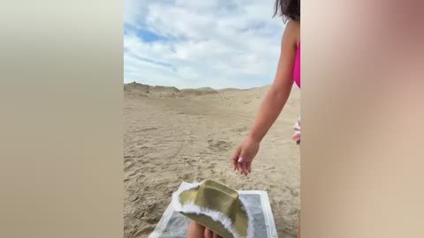 Media: Video of a young person with light skin and long dark hair in a pink tank top, standing on a sandy beach, holding a beige towel with white trim. The sky is cloudy.