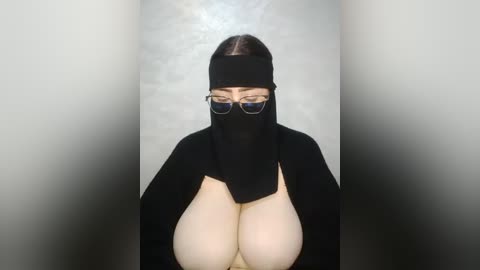 A video of a woman with large, exposed breasts, wearing a black burqa, glasses, and a headband, standing against a gradient grey wall.