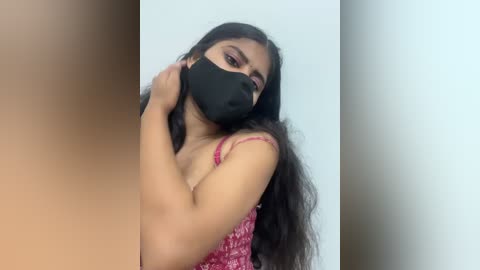Video of a young South Asian woman with long, wavy black hair, wearing a black face mask, pink floral-patterned tank top, and holding her hair. Background is a plain, off-white wall.