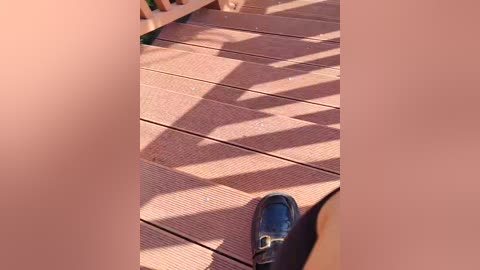 Video of a wooden deck with beige decking planks, featuring geometric shadows cast by a wooden structure. A black shoe with a white sole is centrally positioned.