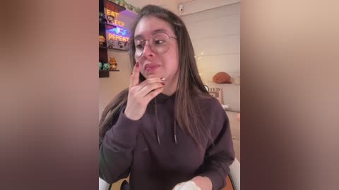 Video of a young woman with long brown hair, wearing glasses, a black hoodie, and a light-colored shirt. She's in a cozy room with colorful posters and a TV, smiling playfully.