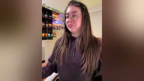 Video of a young woman with long brown hair and glasses, wearing a purple top, sitting in a gaming room with colorful toys and shelves.
