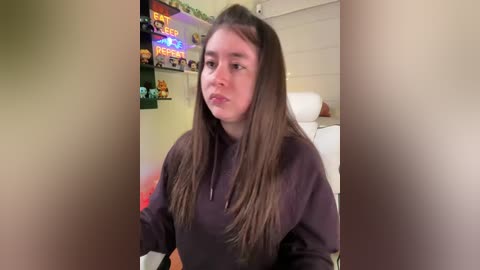 A video of a young woman with long brown hair, wearing a black hoodie, sitting in a brightly lit room with toys, a TV, and a \"FREE STUFF\" sign on the wall.