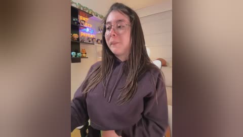 Video of a young woman with long, dark brown hair and glasses, wearing a purple hoodie, standing indoors in a brightly lit room with shelves full of figurines and a TV displaying colorful game titles.