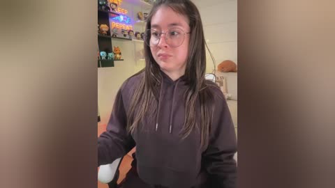 A video of a young woman with long, straight brown hair, wearing glasses, a black hoodie, and a white shirt, sitting in a cluttered room with shelves filled with toys and plushies.