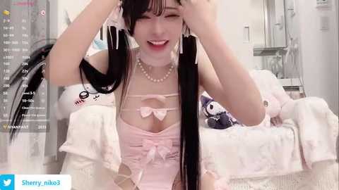 A video of a young Asian woman with long black hair and bangs, wearing a pink lingerie top, adjusting her hair in a room filled with white decor and stuffed toys.