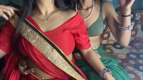 Video of two South Asian women in traditional red and green sarees, adorned with gold embroidery, seated on a patterned cushion.