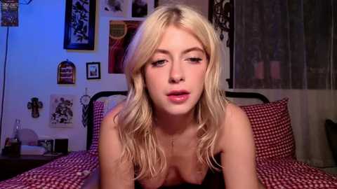 Video of a blonde, fair-skinned woman with medium breasts, wearing a red checkered bra, leaning forward on a bed in a cozy, dimly-lit bedroom with eclectic wall decorations.