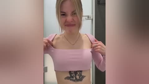 Media: Video of a young blonde woman with fair skin, wearing a cropped pink top, revealing a large black cat tattoo on her ribcage. She stands in a doorway, with blurred faces in the background.