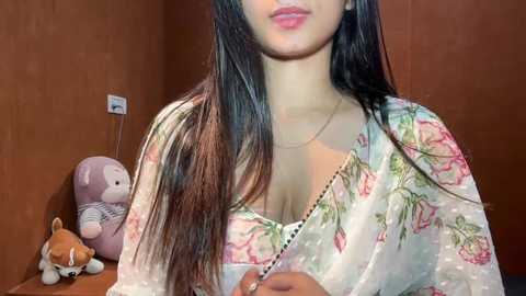 Video of a young woman with long black hair, wearing a floral-patterned blouse, standing in a room with a stuffed bear and a power outlet visible in the background.