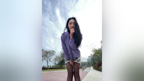 Video of an Asian woman with long black hair, wearing a purple jacket and shorts, standing on a paved path with greenery and a cloudy sky in the background.