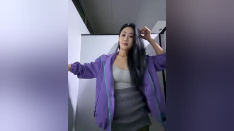 Video of an Asian woman with long black hair, wearing a purple jacket over a grey dress, in a dimly lit hallway.