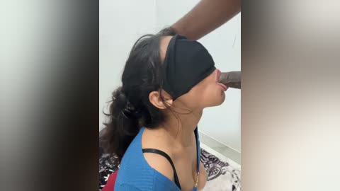 A video shows a young woman with dark hair and a black blindfold kneeling on a patterned rug, her mouth open, receiving oral sex from an unseen man.