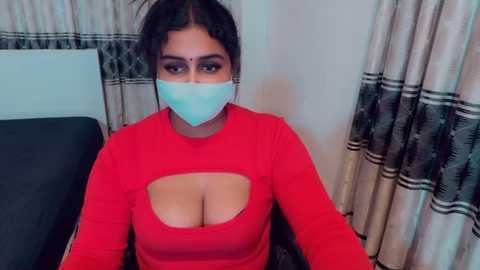 Video of a South Asian woman with medium skin tone, dark hair, and large breasts, wearing a red long-sleeved top with a cutout, a blue face mask, and black curtains in the background.