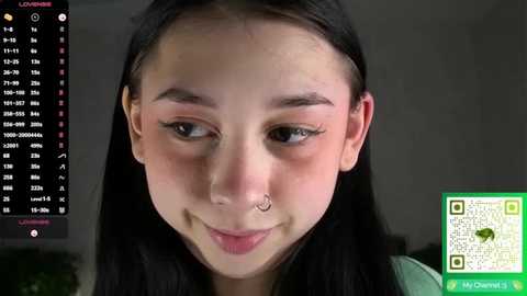 Video of a young woman with long black hair, fair skin, and a nose ring, smiling, indoors. Calendar and QR code in background.