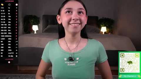 Video of a smiling young woman with straight black hair, wearing a green t-shirt, standing in a dimly lit bedroom with a bed and nightstands.