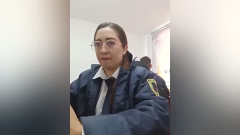 Video of a young woman with fair skin and glasses, wearing a navy blue police uniform with a yellow patch, seated in a dimly lit room.