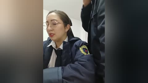 Video of a young Asian woman in a navy-blue police uniform with a badge and glasses, sitting in a dimly lit room with a blurred figure in the background.
