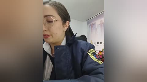 Video of a young woman with long dark hair, wearing glasses, a navy blue police jacket with patches, and a white shirt, sitting indoors with a blurred background.