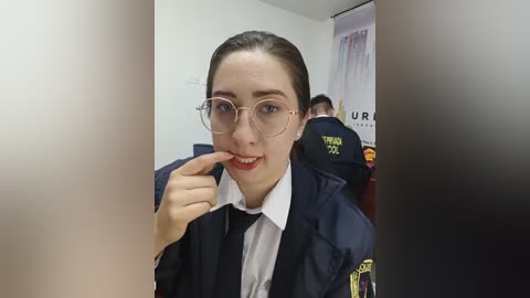 Video of a young woman with light skin, dark hair, and glasses, wearing a navy uniform with a white shirt and name badge, standing in a room with two other officers.