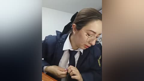 Video of a young Asian woman with light skin, wearing glasses and a navy blue blazer over a white shirt and tie, adjusting her tie in a dimly lit room.