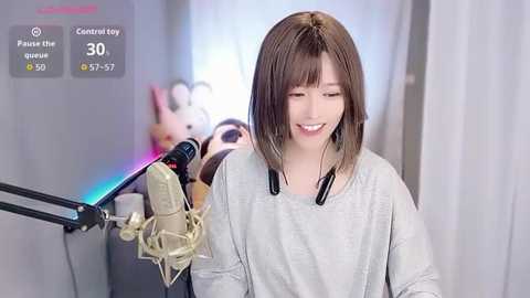 Video of a smiling East Asian woman with shoulder-length brown hair, wearing a grey sweater, singing into a microphone, surrounded by plush toys in a soft, colorful room.