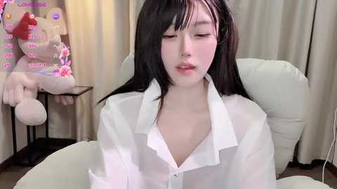 Video of a young East Asian woman with long black hair, wearing a loose white shirt, seated on a white chair. Background features plush toys and beige curtains.