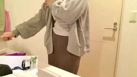 Video of a person in a bathroom, wearing a gray sweater, brown skirt, and a pink towel, standing in front of a beige door, with a white countertop visible.