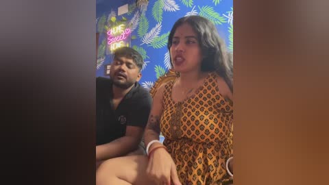 Video of two South Asian people, a man and woman, sitting close. The woman wears a patterned dress, and the man a black T-shirt, in a room with a vibrant blue, leaf-patterned wall and neon sign.