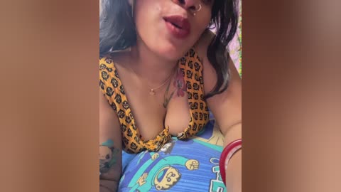 Media: A video of a topless woman with light skin and tattoos, wearing a yellow leopard-print bra, kissing the camera, lying on colorful pillows.