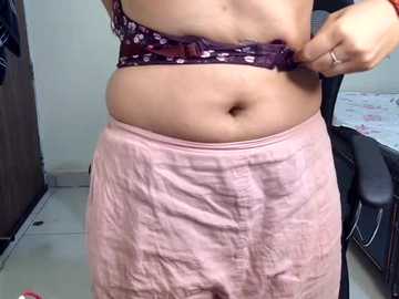 Video of a person with light brown skin, wearing a purple bra with white flowers and a pink skirt, adjusting their bra, in a dimly lit room with a bed and a dark wardrobe visible.