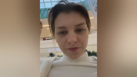 Video of a young Caucasian woman with light skin and dark hair in a bun, wearing a white sweater, standing in a modern indoor setting with large glass windows and greenery.