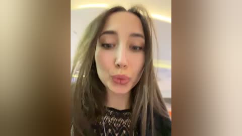 A video of a young woman with long, straight brown hair, light skin, and full lips puckering for a kiss. She wears a black lace top. The background is blurred, showing an indoor setting with a circular ceiling light.