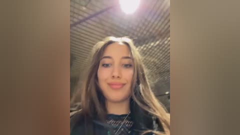 A video of a young woman with long, wavy brown hair and a confident smile, wearing a black top, against a background of a dimly lit, grid-patterned ceiling.