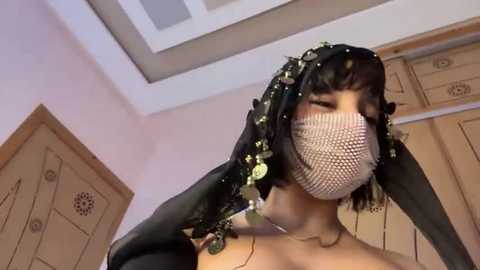 Video of a woman with light skin, wearing a black, beaded veil, fishnet mask, and black gloves, standing indoors against a beige backdrop with wooden doors.