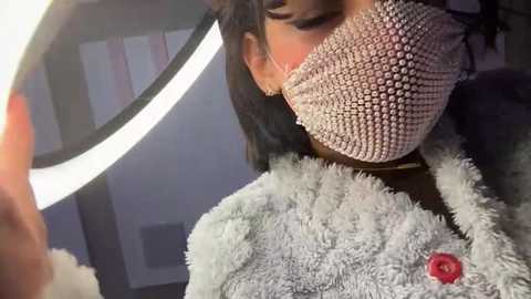 A video of a person wearing a pink, beaded face mask and a fuzzy, light gray sweater with a red flower. Background features a round mirror and dimly lit room with a white lamp.
