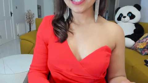 Video of a smiling woman with medium skin tone, wearing a red one-shoulder dress, silver earrings, and a white fluffy pillow on a mustard-yellow couch.