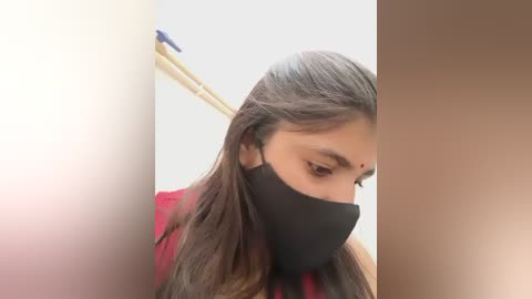 Media: Video of a South Asian woman with long, dark hair, wearing a black face mask and a red shirt, looking down, in a bathroom with a white wall and yellowish towel rack.