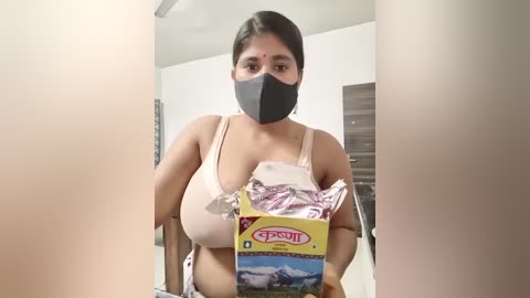 Video of a South Asian woman with medium skin tone and dark hair, wearing a black mask, beige bra, and gray pants, holding a box of Haldiram's sweets in a clean, modern bathroom.