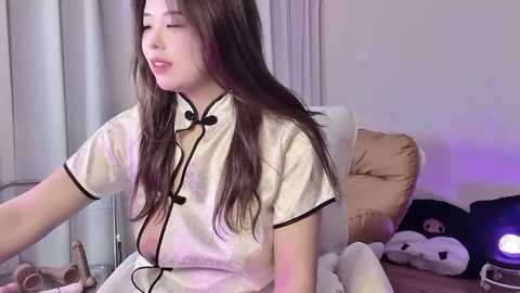 Video of an Asian woman with long brown hair, wearing a white satin robe, sitting on a bed with plush toys, including a panda, in a softly lit room.