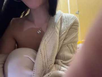 Video of a topless woman with light skin, wearing a beige knitted cardigan, her long black hair partially visible, holding a white bra, in a yellow bathroom.