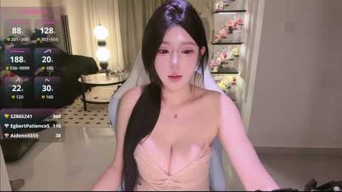 Video of an Asian woman with long black hair in a white strapless dress, sitting on a chair in a dimly lit room with a table, lamp, and shelves in the background.