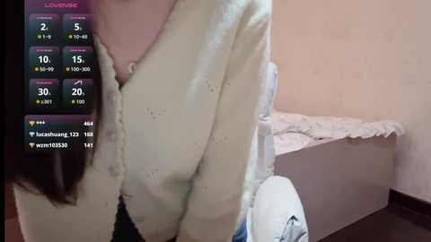 Media: A video showing a woman with a pale complexion, wearing a cream-colored cardigan and a white blouse. She holds a white bag with blue and green patterns. In the background, a bed with white sheets is visible.